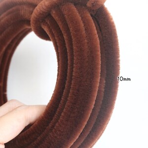 May include: A coiled brown pipe cleaner with a fuzzy, textured surface. The pipe cleaner has a diameter of 10mm as indicated by the text printed next to the coil. The material is a deep, solid brown color with a soft, dense, and fibrous appearance.