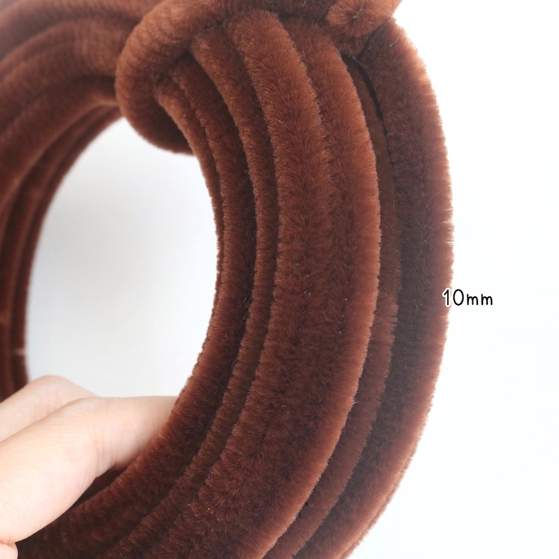 May include: A coiled brown pipe cleaner with a fuzzy, textured surface. The pipe cleaner has a diameter of 10mm as indicated by the text printed next to the coil. The material is a deep, solid brown color with a soft, dense, and fibrous appearance.