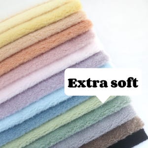 Puede incluir: Una pila de tela suave y esponjosa en varios colores pastel, incluyendo amarillo, melocotón, rosa, lavanda, azul, verde y gris. La tela se describe como "Extra suave".