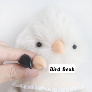 Op de afbeelding: Een wit pluche vogel-speeltje met een zachte, pluizige textuur, kleine zwarte oogjes en een lichtgekleurde stoffen kraag met een geplooide rand. Het speeltje bevat een afneembare, gebogen vogelbek in zowel zwart als licht perzik.