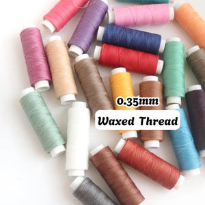 Puede incluir: Una colección de carretes de hilo encerado de 0,35 mm en varios colores, incluyendo rosa, morado, azul, rojo, verde, amarillo, marrón, blanco y naranja. Los carretes están dispuestos de forma dispersa, cada uno con un núcleo de plástico blanco.