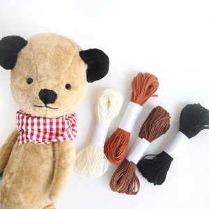 Pode incluir: Um urso de peluche castanho com uma echarpe xadrez vermelha e branca e cinco rolos de fio de bordado em diferentes cores: branco, laranja, castanho, preto e castanho claro.