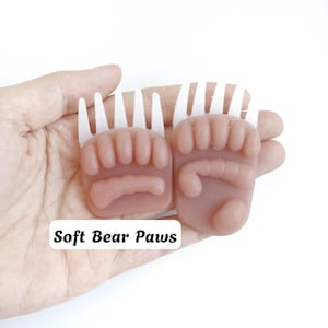May include: Two light brown, semi-translucent bear paw-shaped objects with white, comb-like protrusions at the top. Each piece features molded toe and pad details. The text "Soft Bear Paws" is printed in black on a white rectangular label.