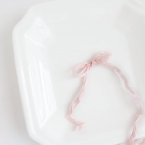 May include: A pink ribbon tied in a bow on a white ceramic plate.