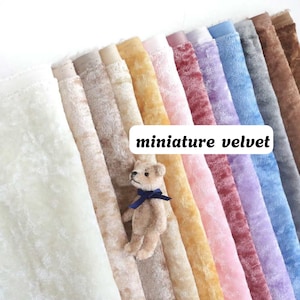 Op de afbeelding: Een collectie miniatuur fluwelen stofstalen gerangschikt in een rij, met kleuren zoals wit, beige, tan, geel, roze, mauve, paars, blauw en bruin. Een kleine, lichtbruine teddybeer met een donkerblauw lint zit op de stof.