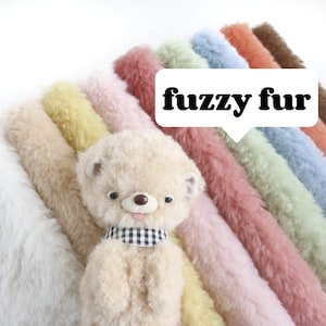 Könnte beinhalten: Eine Auswahl an flauschigen Stoffen in verschiedenen Farben, darunter Weiß, Beige, Gelb, Rosa, Hellblau und Hellgrün. Die Stoffe sind in einem Regenbogenmuster angeordnet, wobei der weiße Stoff unten und der hellgrüne Stoff oben liegt. Der Text "fuzzy fur" ist in schwarzen Buchstaben auf weißem Hintergrund geschrieben.