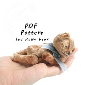 May include: A small, light brown plush bear in a lying-down position, featuring a dark grey ribbon tied around its neck. The text "PDF Pattern lay down bear" is printed in black above the bear. The bear has a textured, fuzzy surface.