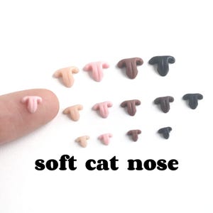 May include: A collection of small, T-shaped cat noses in various colors, including tan, pink, brown, and black. The text "soft cat nose" is displayed in black font below the noses. The noses are arranged on a white surface.