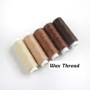Può includere: Cinque rocchetti di filo cerato disposti in una fila diagonale, con colori che sfumano dal crema al beige, marrone chiaro, marrone medio e marrone scuro. Ogni rocchetto ha un nucleo in plastica bianca e il testo Wax Thread è visibile nell'angolo.