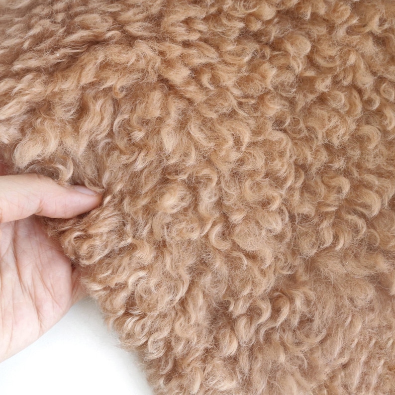 May include: Close-up of a tan-colored, curly faux fur fabric. The texture is dense and plush, with a soft, wavy appearance. The fabric is being held by a hand, showing the flexibility and depth of the material. Ideal for crafting and textile projects.
