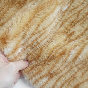 May include: Close-up of a faux fur fabric with a tiger stripe pattern. The fabric features alternating bands of orange and light brown, with a soft, textured appearance. The fur is held by a hand.