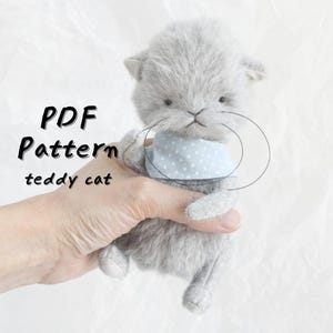 May include: A gray teddy cat with a light blue polka dot scarf. The text "PDF Pattern teddy cat" is written in black on the left side of the image. The cat is held in a person's hand.
