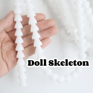 Op de afbeelding: Twee witte plastic poppen skeletten, elk gemaakt van een reeks verbonden kegels, worden in een hand gehouden. De tekst "Doll Skeleton" is onder de skeletten gedrukt.