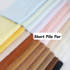 Puede incluir: Una colección de muestras de tela de piel de pelo corto dobladas en una variedad de colores que incluyen azul claro, amarillo, blanco, rosa, beige, naranja, marrón y negro. Se ve una etiqueta rectangular blanca con el texto negro Short Pile Fur.