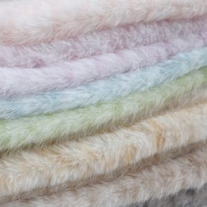 May include: Close-up of a stack of faux fur fabric in various pastel colors. The colors include pink, lavender, light blue, green, beige, and gray. The fur appears soft and fluffy, with a slight sheen.