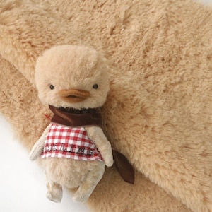May include: A small, plush duckling toy with a red and white checked waistcoat and a brown scarf. The duckling is wearing a brown scarf and has the words "Duckling" embroidered on its waistcoat.