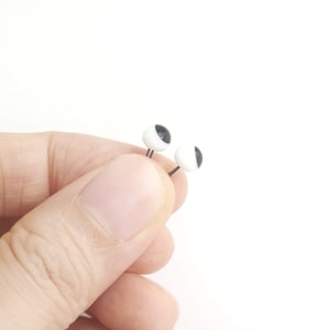 May include: A pair of small, round stud earrings featuring a white sphere with a black circular pupil design, resembling an eye. Each earring has a thin, dark metal post extending from the back of the white and black spherical component.