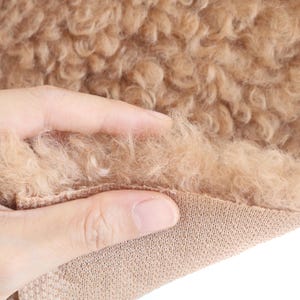 May include: Close-up of a tan, curly faux fur fabric. The fabric is soft and fluffy, with a dense texture. The base of the fabric is a knitted material. The image shows the texture of the faux fur being touched.