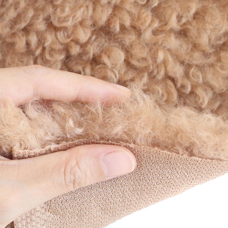 May include: Close-up of a tan, curly faux fur fabric. The fabric is soft and fluffy, with a dense texture. The base of the fabric is a knitted material. The image shows the texture of the faux fur being touched.