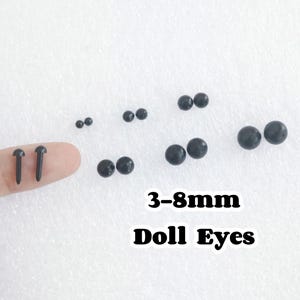Puede incluir: Surtido de ojos de muñeca de plástico negro en varios tamaños, que van de 3 a 8 milímetros. Los ojos están dispuestos en pares sobre una superficie blanca.