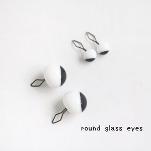 May include: Four round glass eyes with a black and white colour scheme, each featuring a metal wire loop attachment. The eyes are arranged in two pairs of different sizes, and the text round glass eyes is printed in the bottom right corner.