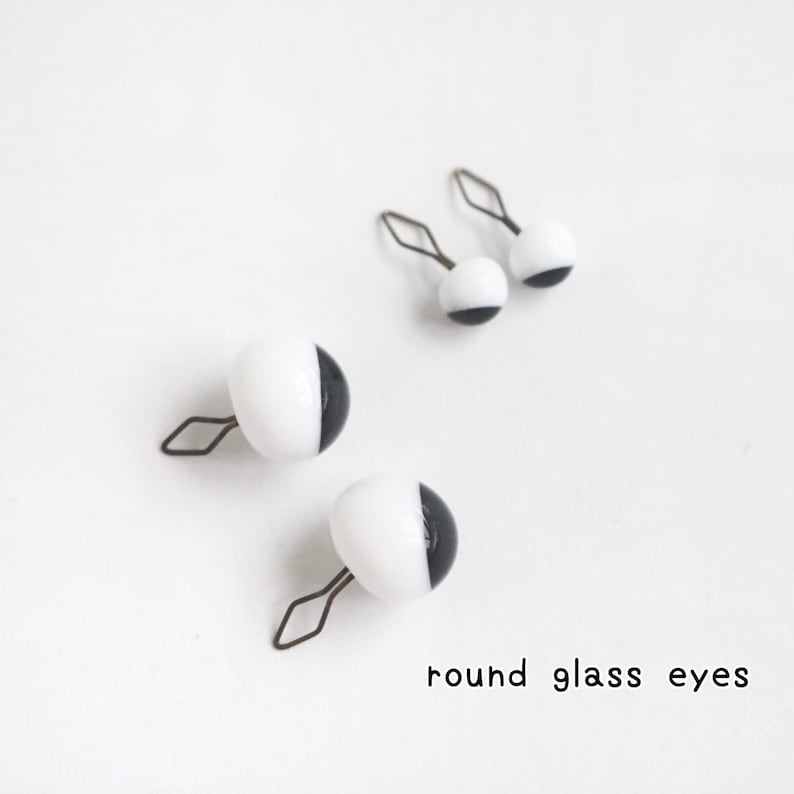 May include: Four round glass eyes with a black and white colour scheme, each featuring a metal wire loop attachment. The eyes are arranged in two pairs of different sizes, and the text round glass eyes is printed in the bottom right corner.