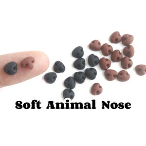 Può includere: Una collezione di piccoli nasi di animali morbidi in nero e marrone. I nasi sono di varie dimensioni e forme, con un design arrotondato. Il testo "Soft Animal Nose" è visualizzato in lettere nere in grassetto.
