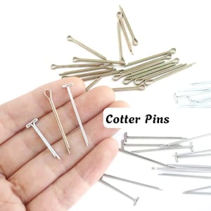 Puede incluir: Una colección de pasadores de chaveta metálicos en tonos plateados y dorados, con un diseño de pasador dividido con cabeza en forma de bucle. Los pasadores están dispersos en grupos, con tres unidades destacadas. El texto Cotter Pins es visible.