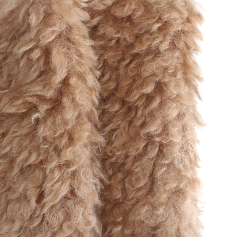 May include: Close-up of a tan, fluffy fabric with a curly texture. The material appears soft and plush, with a dense, shaggy appearance. The fabric is likely used for clothing or home decor.