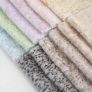 May include: A collection of faux fur fabric samples in various pastel colors, including light blue, green, pink, and beige. The fabrics have a soft, fluffy texture and a spotted pattern, creating a luxurious appearance. Ideal for craft projects or fashion design.