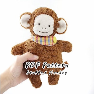 May include: A brown plush monkey toy with a textured, curly fabric body, a smooth white face, and large rounded ears. It wears a colorful plaid neckerchief secured with a yellow button. The text PDF Pattern Stuffed Monkey is visible.