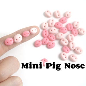 Può includere: Primo piano di una collezione di piccoli bottoni a forma di naso di maiale rosa e bianco. I bottoni sono di forma ovale con due piccoli fori. Il testo "Mini Pig Nose" è visibile nella parte inferiore dell'immagine.