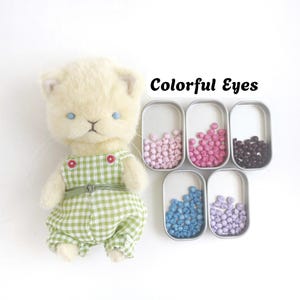 Puede incluir: Una muñeca de gato de peluche color crema con un overol de cuadros verdes y blancos con botones rojos, junto a cinco pequeñas latas de metal que contienen cuentas redondas diminutas en tonos rosa claro, rosa oscuro, marrón oscuro, azul y lila. El texto dice Colorful Eyes.