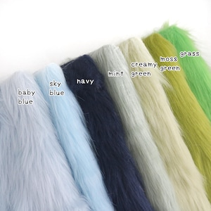 Op de afbeelding: Een collectie kunstbont stofstalen gerangschikt in een rij, met de kleuren gelabeld als baby blue, sky blue, navy, mint, creamy green, moss green en grass. Elke staal toont een lange, zachte en getextureerde pool in de betreffende kleur.