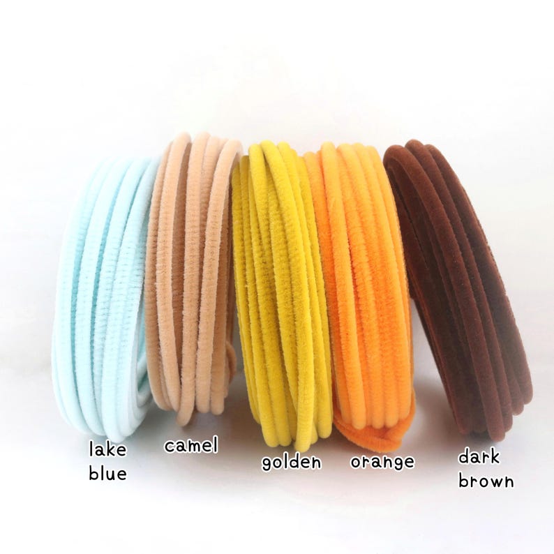 May include: Five bundles of flexible, fuzzy craft stems arranged in a row. The colors from left to right are labeled as lake blue, camel, golden, orange, and dark brown. Each bundle consists of multiple thin, textured, wire-core stems.
