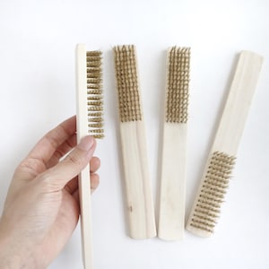 May include: Four rectangular cleaning brushes with light-colored wooden handles and stiff metallic brass-colored wire bristles. Each brush features a long, flat handle and a single row of dense, upright wire bristles designed for scrubbing tasks.