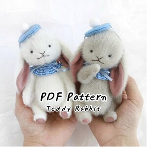 May include: Two small plush rabbit toys with fuzzy white and light gray fur, wearing blue berets with white pom-poms, blue gingham ruffled collars, and pink inner ears. The text PDF Pattern Teddy Rabbit is centered between the two figures.