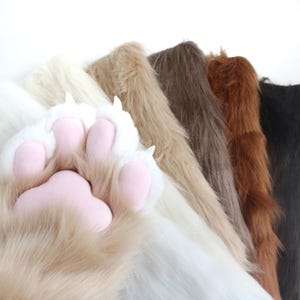 Puede incluir: Primer plano de una pata de peluche con almohadillas rosas y garras blancas, sobre un fondo de varias telas de piel sintética en tonos blanco, beige, marrón y negro. Las telas están dispuestas en diagonal.