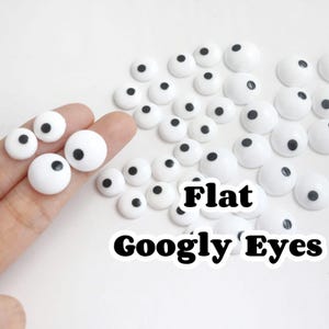 Puede incluir: Una colección de ojos saltones blancos y planos con pupilas negras. La imagen muestra varios tamaños de ojos, algunos sostenidos por una mano. El texto "Flat Googly Eyes" se muestra en blanco y negro.