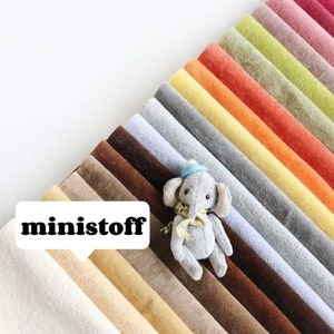 May include: A collection of 20 different colors of soft, plush fabric swatches. The swatches are arranged in a rainbow pattern, with the darkest colors at the bottom and the lightest colors at the top. A small gray plush elephant toy is sitting on top of the swatches.