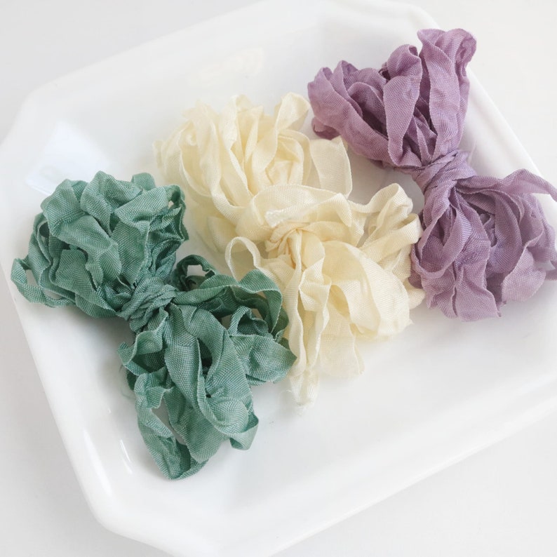 May include: Three spools of ruffled ribbon in green, cream, and purple. The ribbon is arranged in a decorative way on a white plate.