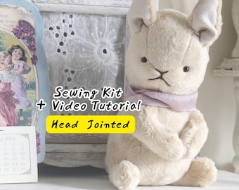 Puede incluir: Un juguete de peluche de conejo color crema con orejas largas y una bufanda morada clara alrededor del cuello. El texto Sewing Kit + Video Tutorial Head Jointed está superpuesto en la imagen. El conejo tiene ojos negros y nariz y boca cosidas.