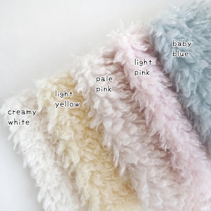 May include: Close-up of soft, textured fabric samples in creamy white, light yellow, pale pink, light pink, and baby blue. Each sample is labeled with its color name. The fabric has a fluffy, curly appearance.