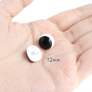 Op de afbeelding: Twee ronde, zwart-witte plastic veiligheidsogen voor knutselwerk, elk met een diameter van 12mm. E&eacute;n oog is geplaatst om de gladde, bolvormige zwarte pupil en witte basis te tonen, terwijl de andere de witte achterkant met een lusje laat zien.