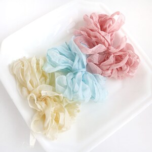 May include: Three spools of ribbon in different colors: light yellow, light blue, and light pink. The ribbon is arranged in a pile on a white surface.