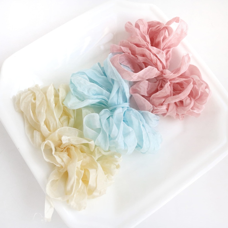 May include: Three spools of ribbon in different colors: light yellow, light blue, and light pink. The ribbon is arranged in a pile on a white surface.