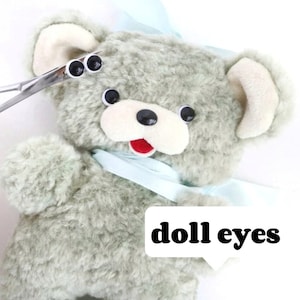Puede incluir: Un oso de peluche gris con una cinta azul alrededor del cuello. El oso tiene ojos negros de botón y una boca roja. El texto "doll eyes" está escrito en letras negras sobre un fondo blanco.