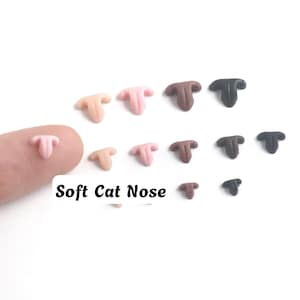 Puede incluir: Una colección de pequeños componentes de nariz de gato suaves en varios tonos de rosa, marrón y negro. Las piezas están dispuestas en filas, con una nariz rosa claro descansando sobre un dedo. El texto Soft Cat Nose está impreso en negro.