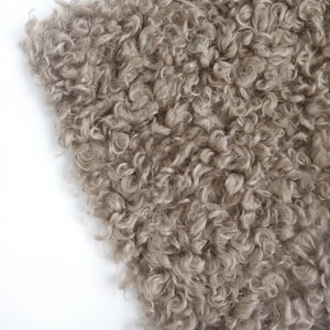 May include: A close-up view of a textured fabric featuring dense, curly, light brown fibers. The material has a shaggy, looped appearance with a consistent pile throughout, creating a soft and voluminous surface with varied light and dark tones.