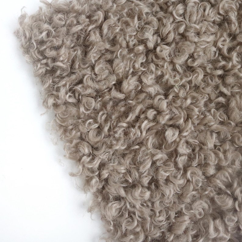 May include: A close-up view of a textured fabric featuring dense, curly, light brown fibers. The material has a shaggy, looped appearance with a consistent pile throughout, creating a soft and voluminous surface with varied light and dark tones.